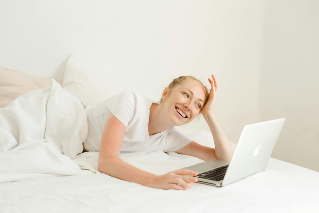 Smiling woman lying on bed using a laptop for leisure or work. Bright, cozy indoor setting.